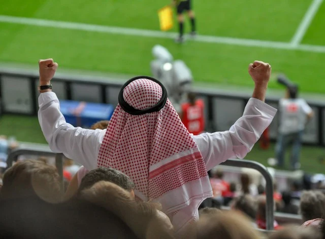 A spectator in traditional Middle Eastern attire raising his fists in celebration at a sports stadium.